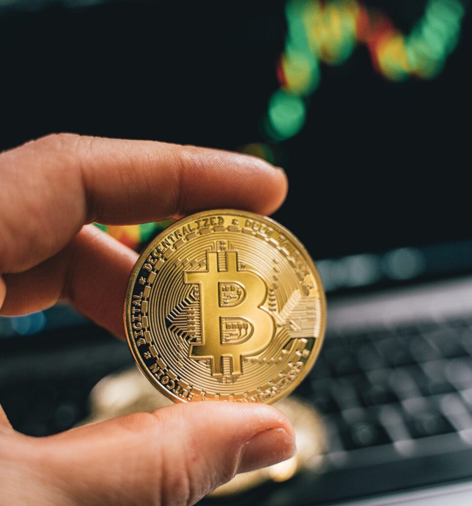 Hand holding a golden Bitcoin against a computer with trading charts, symbolizing finance and cryptocurrency.